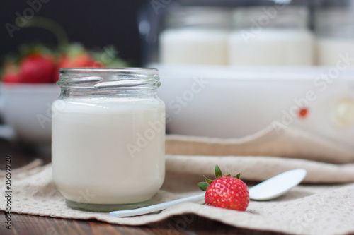 A glass jar of home made yoghurt decorated with no decoration on natural color fabric. One strawberry nearby, more strawberries in blur, a yoghurt maker in blur.