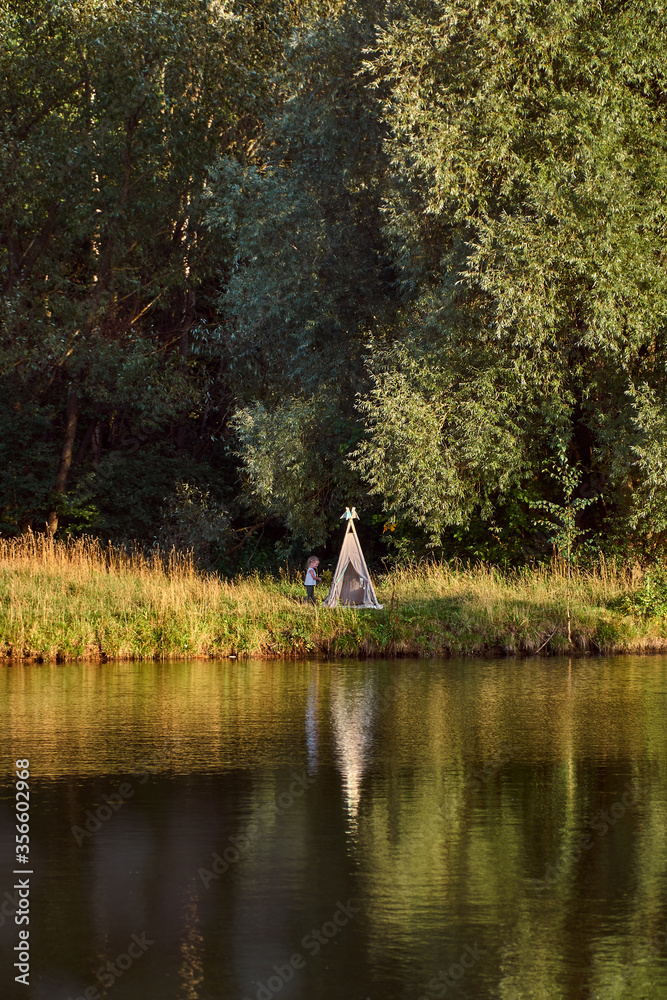 Fototapeta premium Pond on a sunny day. Wigwam on the other shore. Lake in the forest. Green fairy tale. High quality photo