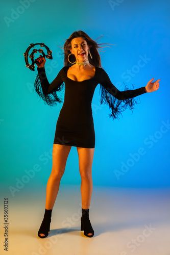 Caucasian female singer with tambourin isolated on blue studio background in neon light. Beautiful female model in black dress. Concept of human emotions, facial expression, ad, music, art, festival.