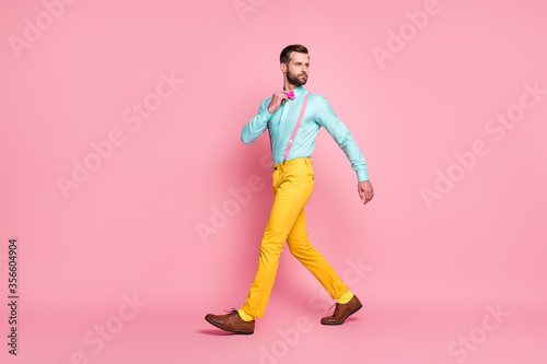 Full length profile photo of handsome guy trend clothes walking office corporate meeting wear shirt suspenders bow tie yellow pants footwear isolated pastel pink color background