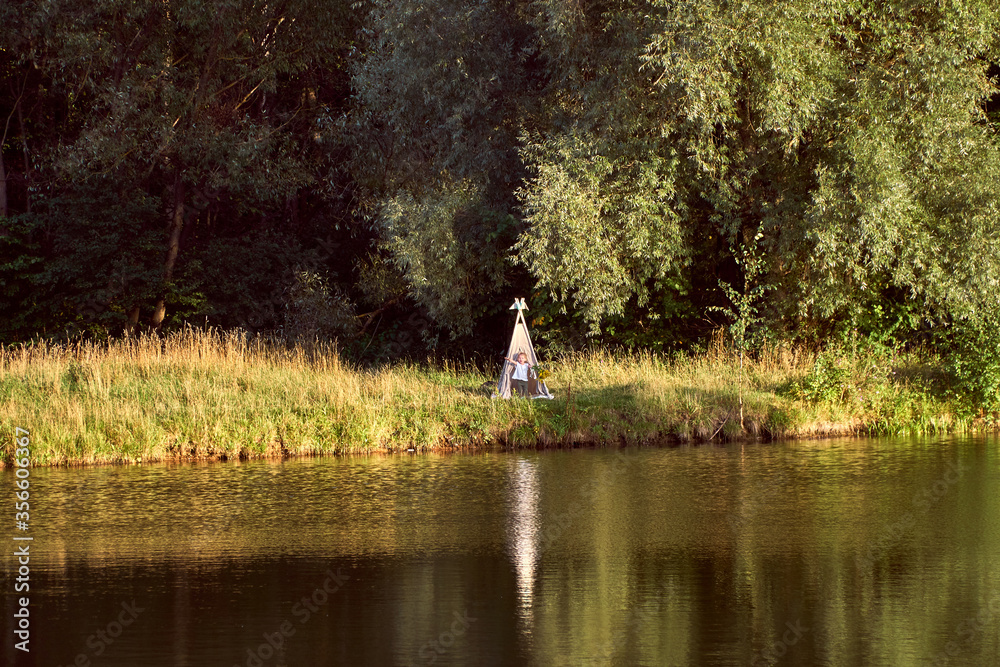 Fototapeta premium Pond on a sunny day. Wigwam on the other shore. Lake in the forest. Green fairy tale. High quality photo