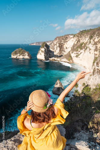 Young happy woman traveller sitting on top of cliff and looking to Diamond beach in a morning. A woman enjoying, exciting and cheer up. Nusa Penida near Bali island, Indonesia