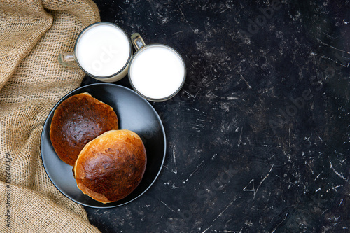 Two homemade buns on a black plate, two glasses of milk and a burlap cloth on a black texture background. Flat lay, studio shot, copy space. Organic healthy breakfast concept.