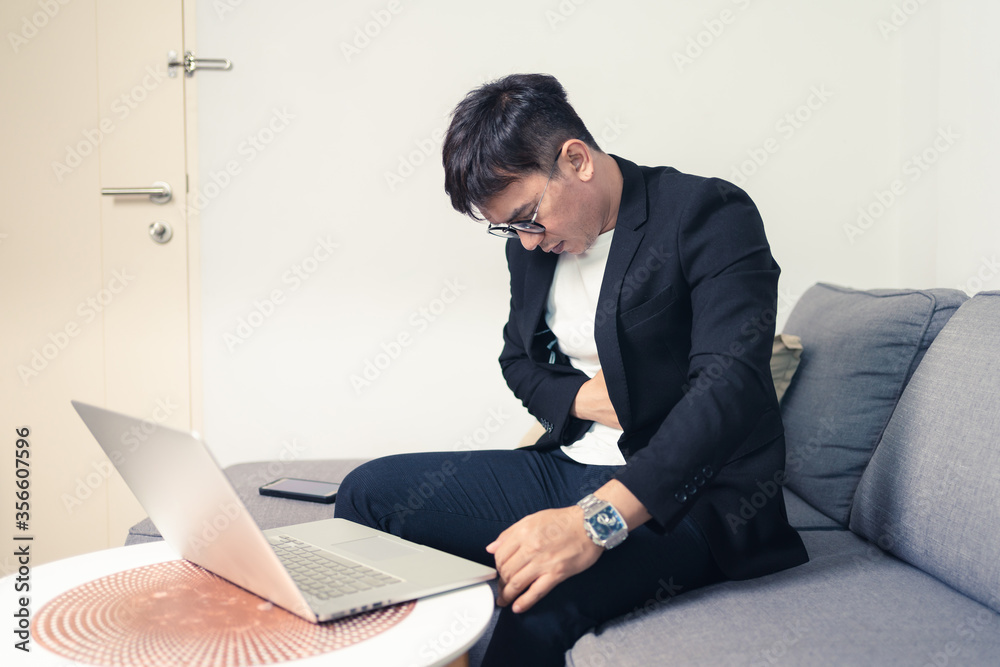 Young Asian business man sitting on sofa and working from home. Man feeling stress and suffering from stomachache. Tired young entrepreneur manager. Work from home concept Young Asian business man sitting on sofa and working from home. Man feeling stress and suffering from stomachache. Tired young entrepreneur manager. Work from home concept