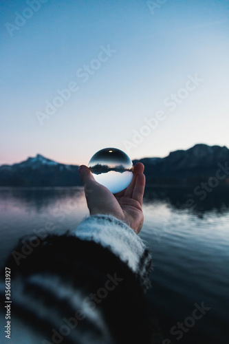 man holding a glassball in front of background with mountains and a lake.