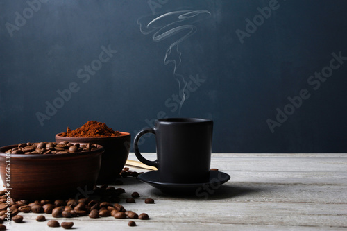 Hot espresso cup with steam, roasted Arabica beans in clay bowls, coffee powder on while table against dark blue wall. Side view, copy space. Coffee shop, morning, baristas workplace concept