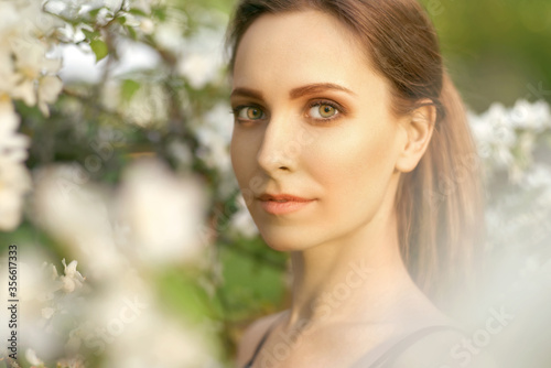 A young smiling girl with green eyes stands in a blooming Apple tree in the spring and sniffs the flowers on a pleasant Sunny day. Close up portrait of a beautiful woman in the Park.