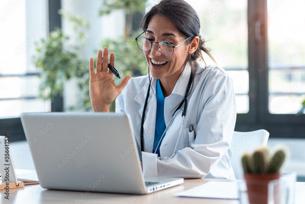 Female doctor waving and talking with colleagues through a video call with a laptop in the consultation. Female doctor waving and talking with colleagues through a video call with a laptop in the consultation.
