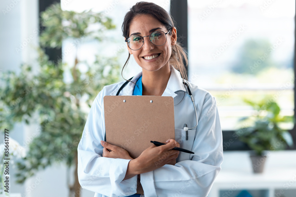 Fototapeta premium Beautiful female doctor looking at camera and smiling while holding medical documents and standing in the consultation.