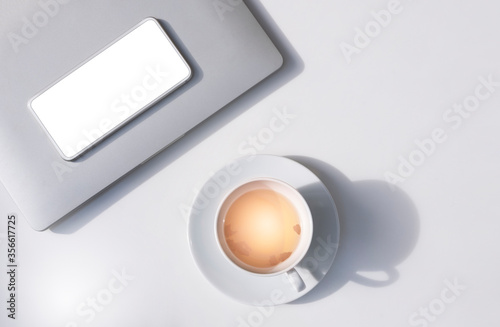 Sunlight and shadow on surface of herbal hot tea in ceramic tea cup with white smartphone and grey laptop on white tabletop