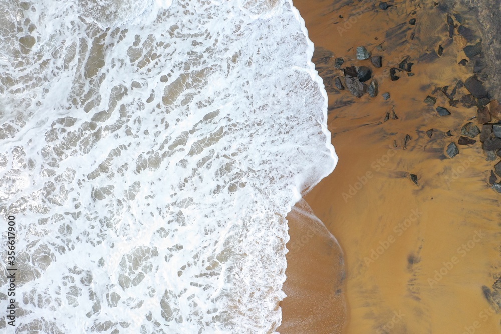 aerial drone bird view shot of the sea shore with yellow sand, black rocks, large white waves and foam crashing on the beach forming beautiful textures, patterns, shapes. Sri Lanka aerial drone bird view shot of the sea shore with yellow sand, black rocks, large white waves and foam crashing on the beach forming beautiful textures, patterns, shapes. Sri Lanka