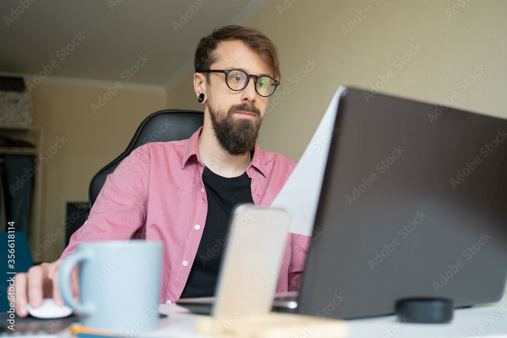 Bearded young man in eyeglasses working at home. Dark-haired attractive guy looking at laptop screen and sitting on isolation. Workplace and freelance concept Bearded young man in eyeglasses working at home. Dark-haired attractive guy looking at laptop screen and sitting on isolation. Workplace and freelance concept