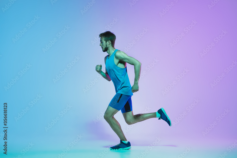 Portrait of active young caucasian man running, jogging on gradient studio background in neon light. Professional sportsman training in action and motion. Sport, wellness, activity, vitality concept.