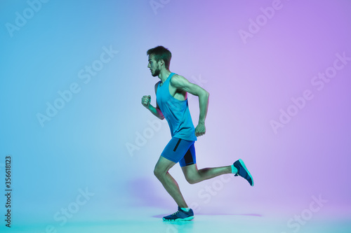 Portrait of active young caucasian man running, jogging on gradient studio background in neon light. Professional sportsman training in action and motion. Sport, wellness, activity, vitality concept.