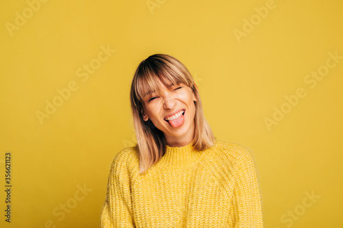 Positive funny playful young blonde woman in yellow sweater posing on camera. Girl showes tongue and have fun alone in studio. Isolated over yellow background. © Anton
