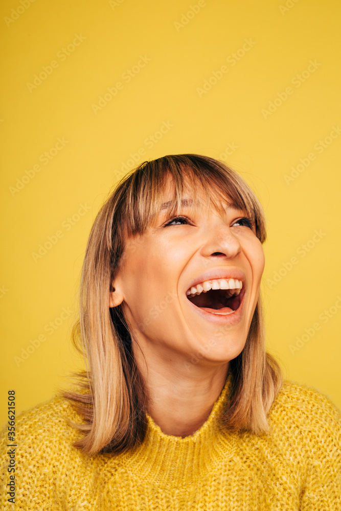 Portrait of cheerful positive young blonde woman laughing out loud and look up. Posing on camera. Vertical picture of woman in yellow sweater isolated over background.