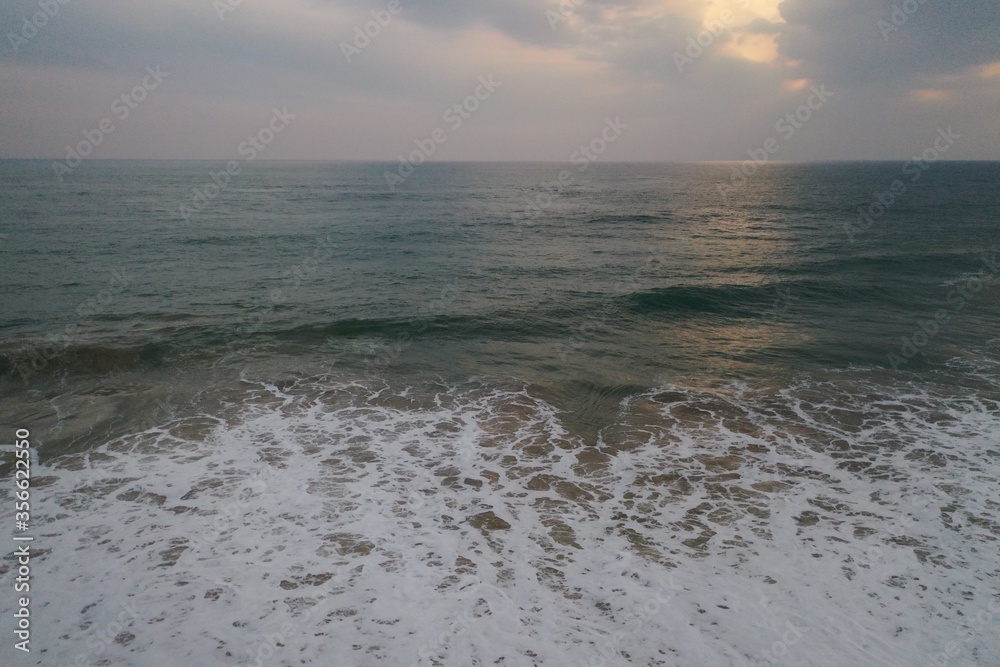 Obraz premium aerial sunset drone bird view shot of the sea surface with turquoise blue water, large white waves and foam forming beautiful textures, patterns, shapes and a cloudy sky on the horizon. Sri Lanka