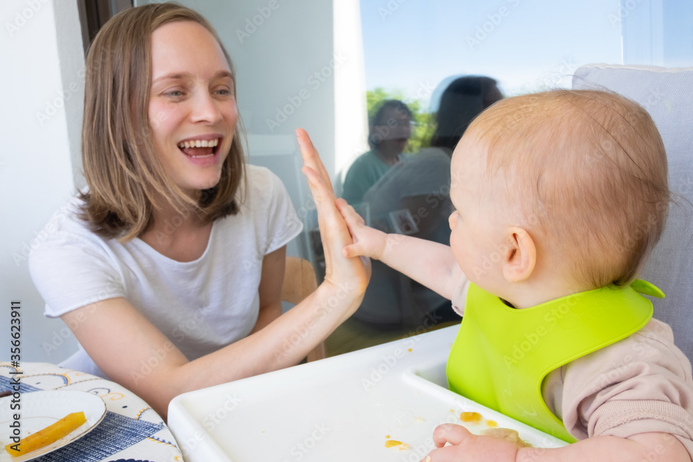 Happy mom and baby daughter giving high five to each other while having family lunch or dinner on balcony. First solid food or child care at home concept Happy mom and baby daughter giving high five to each other while having family lunch or dinner on balcony. First solid food or child care at home concept