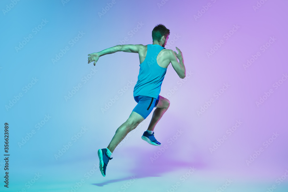 In jump. Portrait of young caucasian man running, jogging on gradient studio background in neon light. Professional sportsman training in action and motion. Sport, wellness, activity, vitality concept