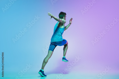 In jump. Portrait of young caucasian man running  jogging on gradient studio background in neon light. Professional sportsman training in action and motion. Sport  wellness  activity  vitality concept