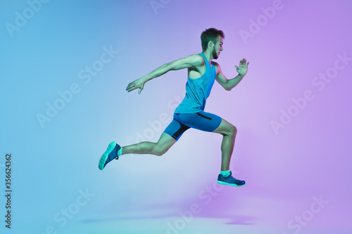 Fototapeta Naklejka Na Ścianę i Meble -  In jump. Portrait of young caucasian man running, jogging on gradient studio background in neon light. Professional sportsman training in action and motion. Sport, wellness, activity, vitality concept
