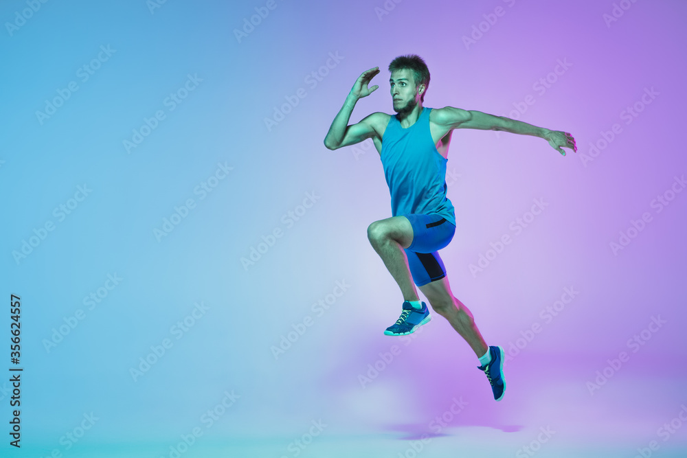 In jump. Portrait of young caucasian man running, jogging on gradient studio background in neon light. Professional sportsman training in action and motion. Sport, wellness, activity, vitality concept