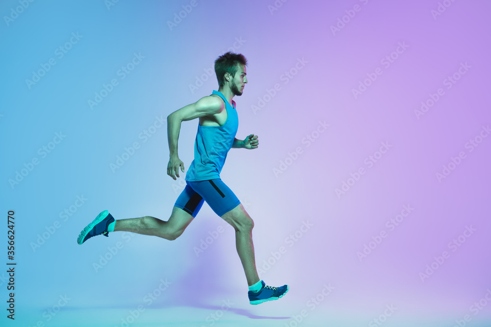 In jump. Portrait of young caucasian man running, jogging on gradient studio background in neon light. Professional sportsman training in action and motion. Sport, wellness, activity, vitality concept