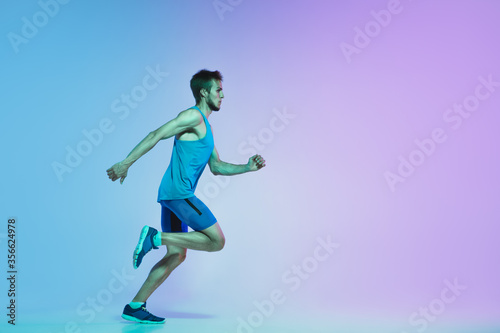 In jump. Portrait of young caucasian man running  jogging on gradient studio background in neon light. Professional sportsman training in action and motion. Sport  wellness  activity  vitality concept