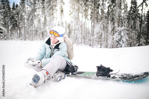 Caucasian woman snowboarder sitting in snow near snowboard on beautiful snowy forest background in sunny day.