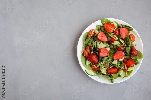 Plate of fresh vegetarian salad with strawberry, spinach, cucumber on the gray background. Copy space.