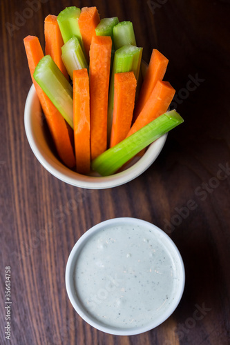 vertical close up isolated top view shot of a bowl of party snack in form of orange carrot and green cerely sticks with a white cup of blue cheese dipping sauce on a dark brown wooden table