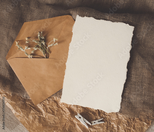 Cerastium dry flower, white empty paper sheet, envelope on natural brown craft sackcloth fabric and crumpled paper flat layout flat-lay top view. Design template for card, invitation, wedding decor