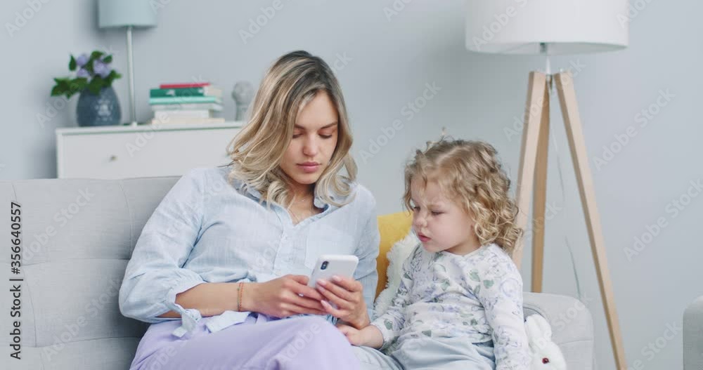 Middle plan of cute daughter having fun with mummy enjoy using modern gadget smart phone playing online games. Happy mom together with little child. Concept of family, parenthood, togetherness. Middle plan of cute daughter having fun with mummy enjoy using modern gadget smart phone playing online games. Happy mom together with little child. Concept of family, parenthood, togetherness.