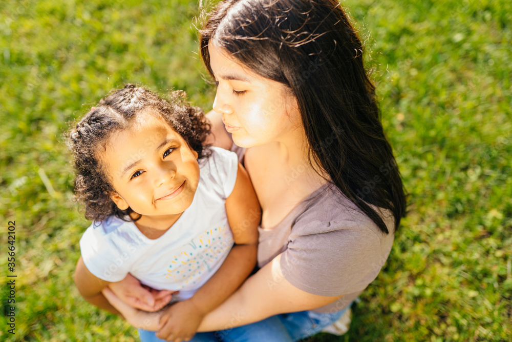 Top view portrait of ecuadorian single mother having funny conversation with her cute curly dark skinned hispanic daughter sits on green grass meadow in sunny summer day, outdoors. Top view portrait of ecuadorian single mother having funny conversation with her cute curly dark skinned hispanic daughter sits on green grass meadow in sunny summer day, outdoors.