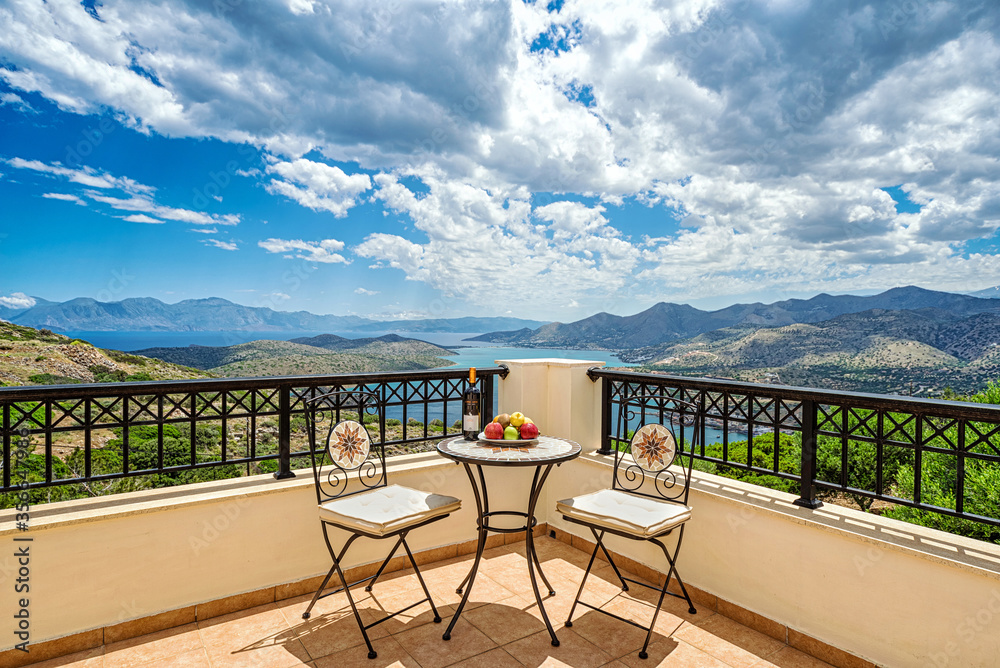 Table and two chairs in the corner of the balcony/terrace with amazing panoramic view to Crete island, Mediterranean sea and blue sky with white clouds. Fruits and bottle of wine located on the table.