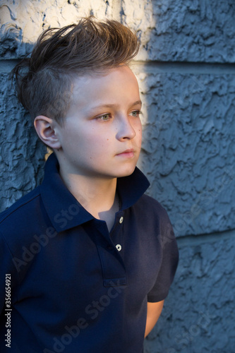 A handsome boy with a fashion haircut is standing by the wall in the street. He is 13 years old.  © IvSky