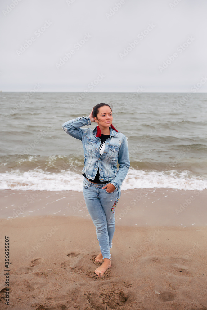 Feminine girl walks along the sand beach near the sea with waves in earrings and hair in the wind in stylish clothes. Good weather. Clouds in the sky. Feminine girl walks along the sand beach near the sea with waves in earrings and hair in the wind in stylish clothes. Good weather. Clouds in the sky.