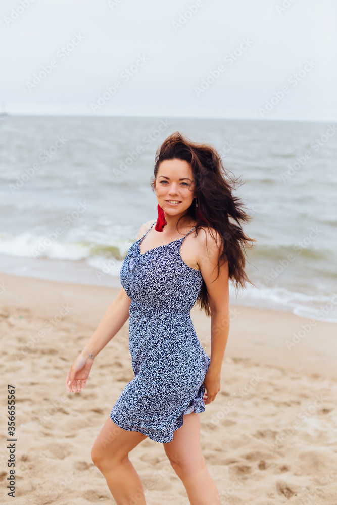 Feminine girl walks along the sand beach near the sea with waves in earrings and hair in the wind in stylish clothes. Good weather. Clouds in the sky. Feminine girl walks along the sand beach near the sea with waves in earrings and hair in the wind in stylish clothes. Good weather. Clouds in the sky.