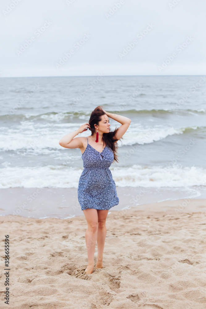 Feminine girl walks along the sand beach near the sea with waves in earrings and hair in the wind in stylish clothes. Good weather. Clouds in the sky. Feminine girl walks along the sand beach near the sea with waves in earrings and hair in the wind in stylish clothes. Good weather. Clouds in the sky.