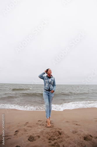 Feminine girl walks along the sand beach near the sea with waves in earrings and hair in the wind in stylish clothes. Good weather. Clouds in the sky.