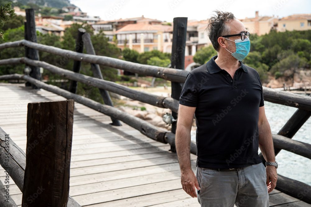 Uomo con mascherina facciale èmaglia nera sta passando sopra un ponte di legno di una località turistica di mare