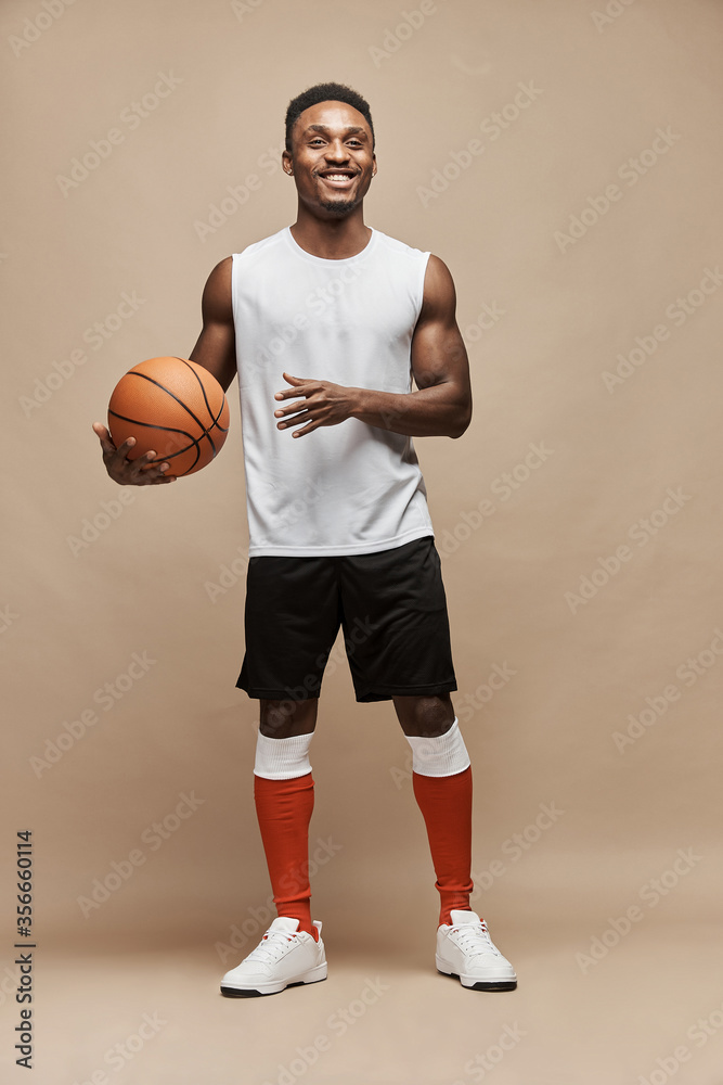 Fototapeta premium full length photo of a black athletic basketball player in the studio on a beige background wearing white T-shirt, black shorts, red long socks and white sneakers, he holds the ball and smiles