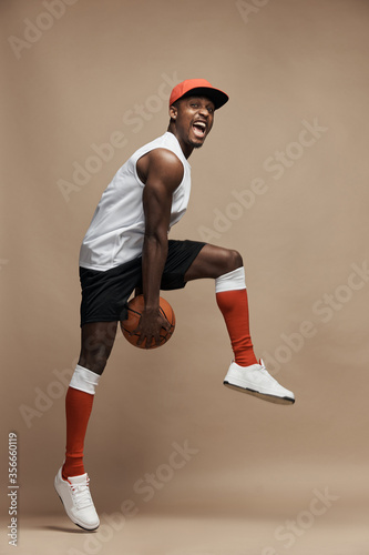 Fototapeta Naklejka Na Ścianę i Meble -  full-length photo of a black  athletic basketball player in the studio on a beige background, jumping high with a ball, wearing white T-shirt, black shorts, red long socks and a cap and white sneakers
