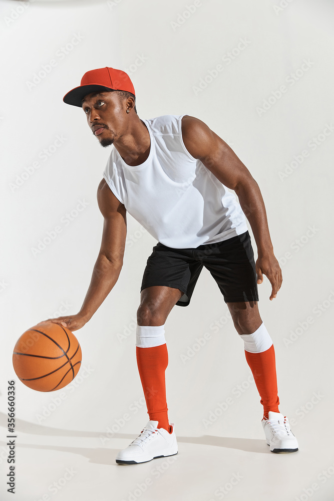 full length photo of a dark-skinned athletic basketball player in studio on a white background posing with a ball, wearing a white t-shirt, black shorts, red long socks and a cap and white sneakers