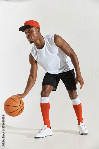 full length photo of a dark-skinned athletic basketball player in studio on a white background posing with a ball, wearing a white t-shirt, black shorts, red long socks and a cap and white sneakers © monchak