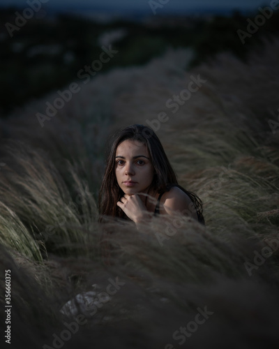 Young woman with long black hair standing between plants looking at the camera during night time after the sunsets with a light hitting her face