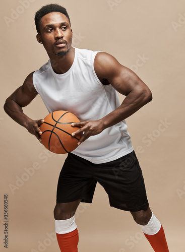 dark-skinned athletic basketball player in studio on a beige background posing with a ball wearing a white T-shirt, black shorts, red long socks © monchak