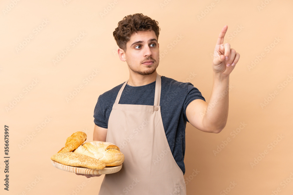 Male baker holding a table with several breads isolated on beige background touching on transparent screen