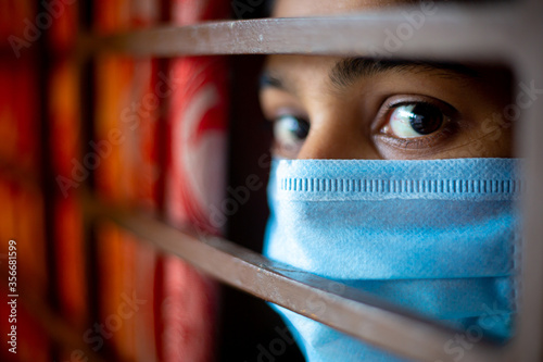 A bored Asian young girl wearing a protection surgical face mask at looking through window being in-home quarantine during coronavirus outbreak. Close-up views. photo
