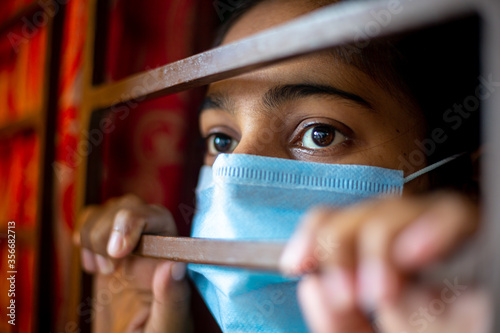 A bored Asian young girl wearing a protection surgical face mask at looking through window being in-home quarantine during coronavirus outbreak. Close-up views. photo